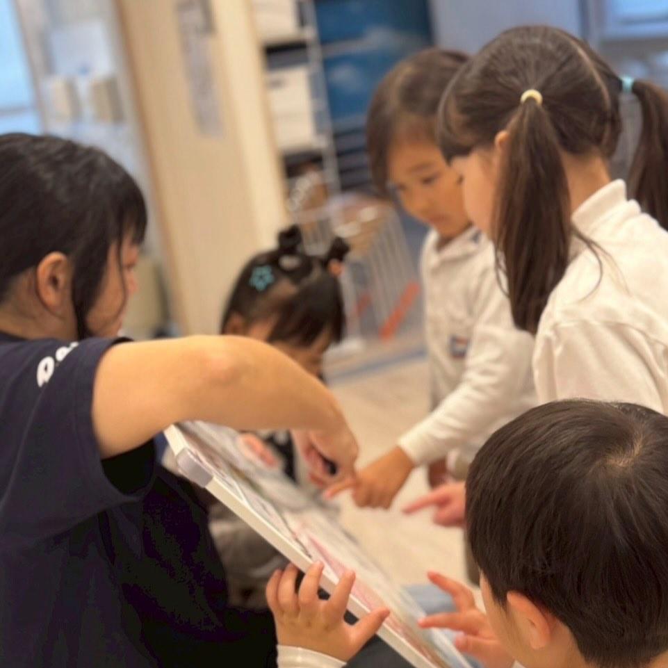 大型絵本を囲んで指をさしながら話し合う園児たちと先生。問いから学びが広がるRaJA Global Academyの探究の時間